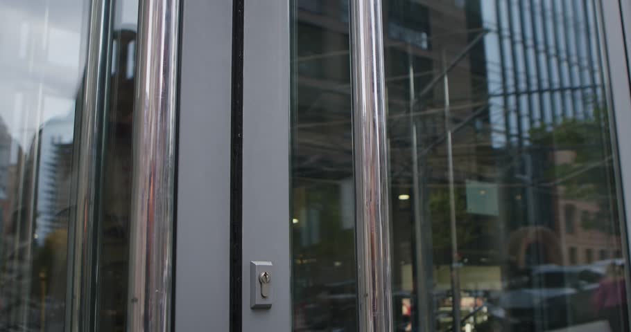 A woman wearing a trench coat opens a glass door, entering a modern building in an urban environment. Slow motion.