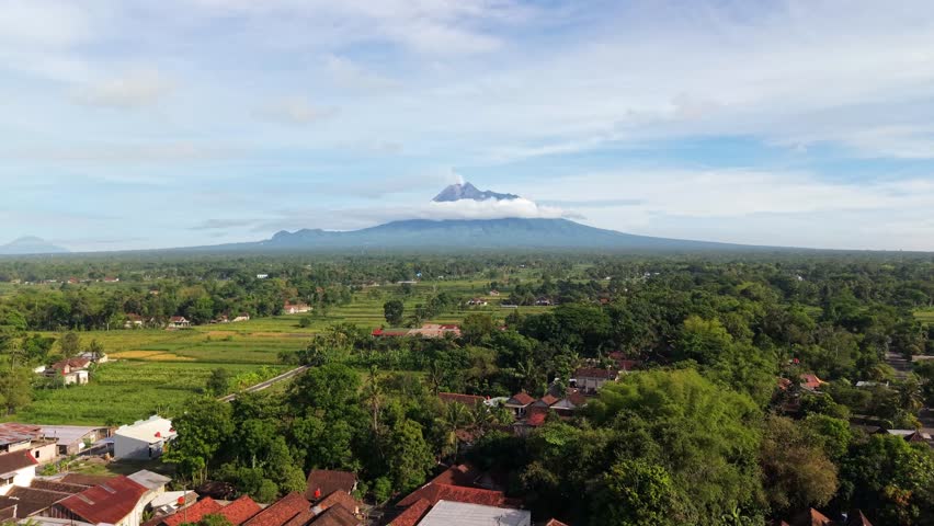 Aerial view of the vast green rice fields contrasting with the majestic Mount Merapi shrouded in white clouds, Klaten Regency, Central Java, Indonesia.