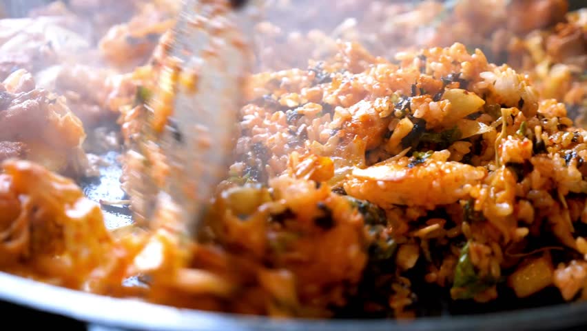 Close up of spatulas stir frying hot steaming Korean fried rice on a large skillet. This spicy Dak Galbi dish includes chicken, vegetables, and seaweed.