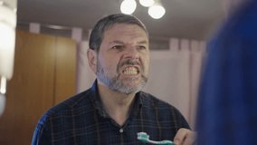 Middle aged man with a beard performing his daily dental hygiene routine, carefully brushing his teeth with a toothbrush and toothpaste while looking at his reflection in the bathroom mirror - Powered by Shutterstock - Get 15% off with code: PIKWIZARD15