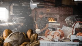 Artisan baker kneads bread dough on a wooden table inside a rustic, smoky bakery. Warm firelight glows from the wood-fired oven as baskets of fresh bread fill the foreground.  - Powered by Shutterstock - Get 15% off with code: PIKWIZARD15