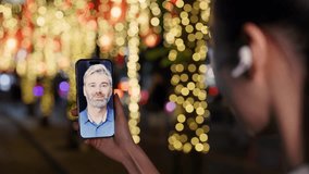 Woman holding smartphone during video call with mature man greeting and smiling under festive Christmas lights in night city. Warm communication, friendship, digital connection and holiday mood. - Powered by Shutterstock - Get 15% off with code: PIKWIZARD15