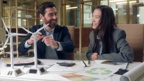 Happy joy Caucasian businessman Korean businesswoman discuss wind turbine model pointing finger laugh talk eco energy office startup sustainable development meeting cooperation innovation paper office - Powered by Shutterstock - Get 15% off with code: PIKWIZARD15