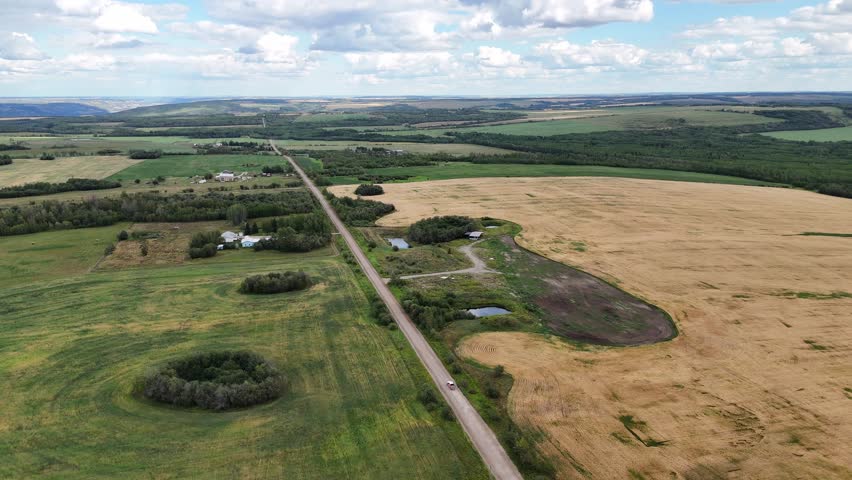 An expansive aerial perspective captures a vast rural expanse, showcasing golden fields ready for harvest, lush green pastures, and a long, straight road cutting through the landscape in Canada