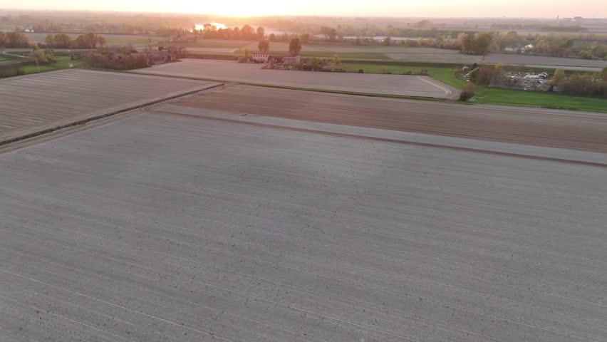 Drone perspective highlights extensive plowed farmland illuminated by golden sunset; winding field borders, green strips, and distant buildings are bathed in evening light, River Po in background.