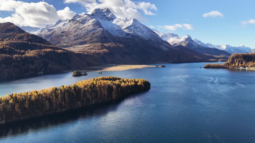 An aerial panorama of Lake Sils (Silsersee) in the Upper Engadin, Switzerland. The deep blue water is surrounded by golden larch forests and snow-capped mountains in a striking autumn display.