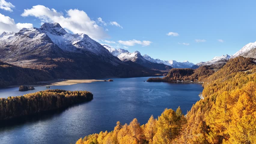 A vast aerial panorama of Lake Silvaplana in the Upper Engadin, Switzerland. Deep blue lake water contrasts with a large golden larch peninsula and a background of massive, snow-capped mountains