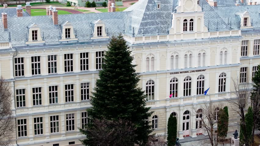 Aerial shot of historic facade of Liviu Rebreanu National College in Bistrita, Romania. The medium shot features a prominent entrance, tall windows, and large evergreen trees flanking the building