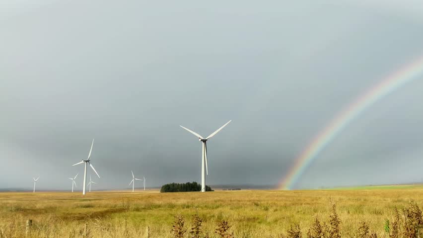  A dramatic drone shot in Caithness, Scotland, captures towering wind turbines standing before a vibrant rainbow against a backdrop of dark, moody skies.