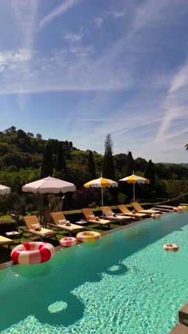 Drone view of a chic villa terrace in Tuscany with bright pools, yellow-white towels on sun loungers, vintage-style inflatable rings, umbrellas, and a Dolce Vita after-wedding party vibe