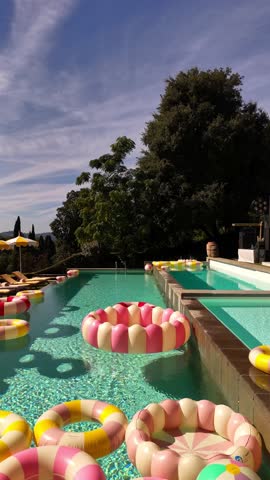 Drone view of a chic villa terrace in Tuscany with bright pools, vintage-style inflatable rings, sun loungers, umbrellas, and a Dolce Vita after-wedding party atmosphere