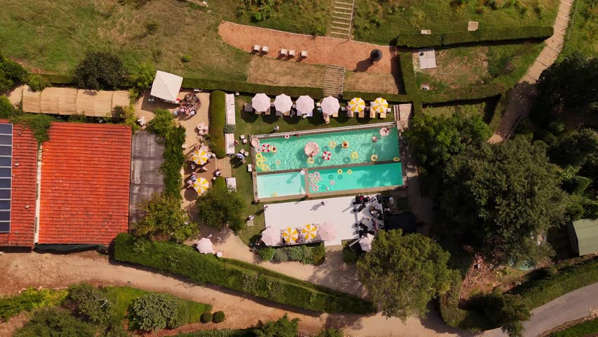 Drone view of a Tuscan villa with red-tiled roofs, bright pools, vintage-style inflatable rings, sun loungers with umbrellas, surrounded by green lawns, creating a Dolce Vita after-wedding party vibe