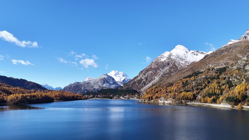 A wide, low aerial view of Lake Silvaplana in the Upper Engadin, Schweiz. The deep blue water reflects a brilliant sky, framed by golden larch slopes and majestic snow-capped mountains.