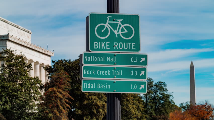Timelapse of a bike route sign near the Lincoln Memorial in Washington, D.C. The sign directs to the National Mall, Rock Creek Trail, and Tidal Basin, with the Washington Monument in the background.