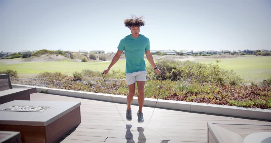 Man holding rope and starting rhythmic jump-rope routine on wooden deck, practicing fitness. Outdoor, cardio, calisthenics, patio, sunlight, silhouette, suburban