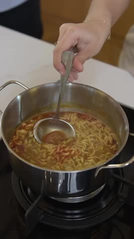 A person stirring instant noodles in a pot on a stove. The hot soup with steam and vibrant broth creates a cozy homemade meal atmosphere in a modern kitchen.