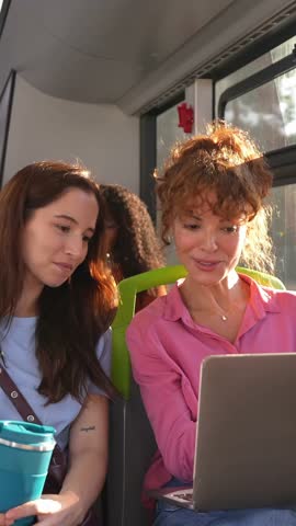Two women commuting together on a public bus. One is working on a laptop while her colleague watches, discussing their work