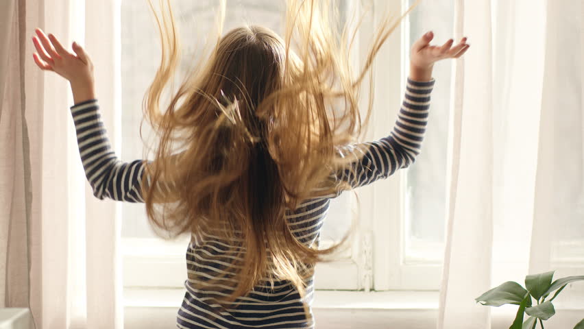 Back view Caucasian little girl child kid looking out window sunny weather shaking long smooth healthy hair turning around smiling going looking at camera walking indoors at home daughter offspring
