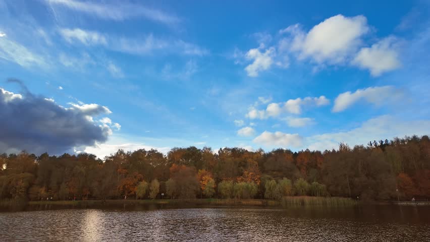 Dramatic clouds contrast with the bright sky above a serene autumn forest by the lake.