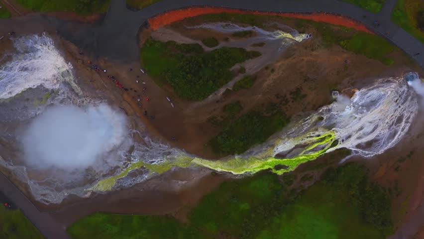 Aerial top down view shows Strokkur hot spring in Haukadalur, Iceland. Steam surges, water flows through yellow green channels, visitors stand on paths and platforms.