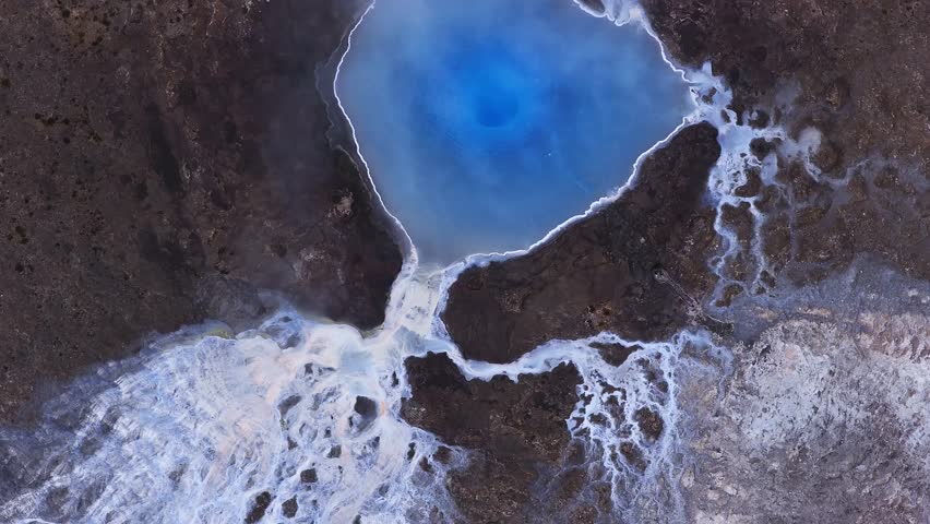 Aerial top down view of a circular deep blue geothermal pool in Iceland, white mineral rings, pale runoff over dark lava rock, thin steam, small blue shelter visible