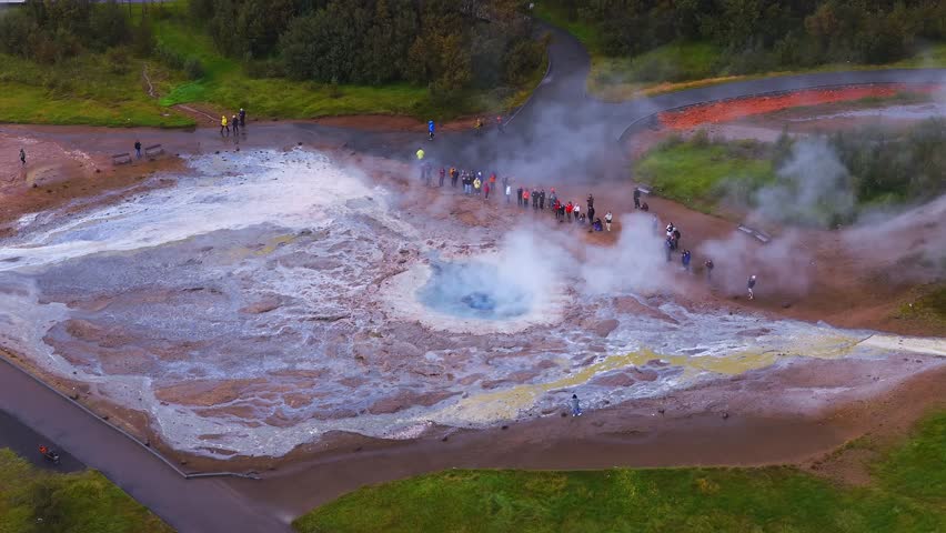 Aerial high angle shot shows a blue geothermal pool at Geysir, Iceland. Steam rises as tourists stand on a curved boardwalk near white terraces and yellow sulfur.