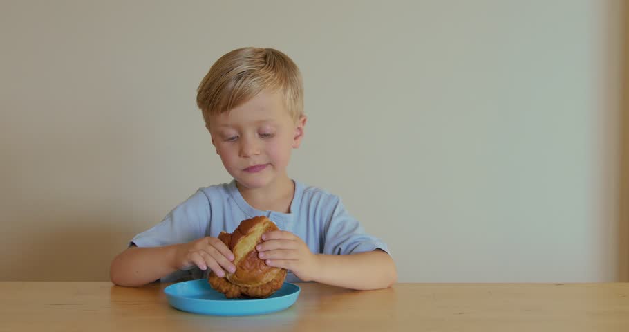 A six-year-old blond boy is eating a chicken burger from a blue plate.