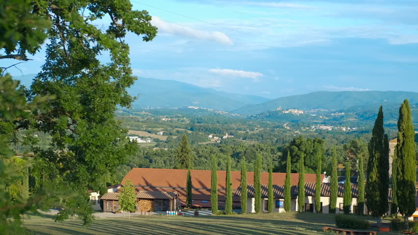 Farmhouse with cypress trees overlooking a valley in the italian countryside. Sunlight illuminates a farmhouse with a row of cypress trees, offering a serene view of the valley and distant mountains
