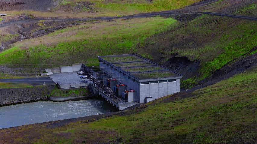 Aerial drone view shows a small hydro plant in Iceland, green roofs, red transformers, spillway, parking with cars, power lines, concrete platform, winding road, daylight
