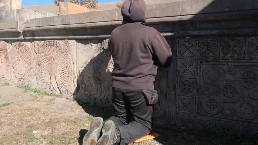 A Christian man kneels by a stone pedestal with ancient symbols and prays making the sign of the cross. A ruined Armenian church is a holy site attracting believers.