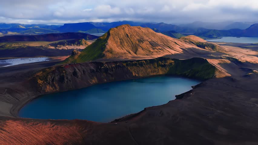 Aerial camera glides above Ljotipollur crater near Landmannalaugar, Iceland. Turquoise lake, rust ridges, black lava fields, distant mountains, soft light, saturated color.