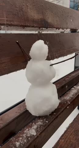 A charming small snowman sits on a wet wooden bench. Tiny sticks serve as arms in this peaceful winter scene.