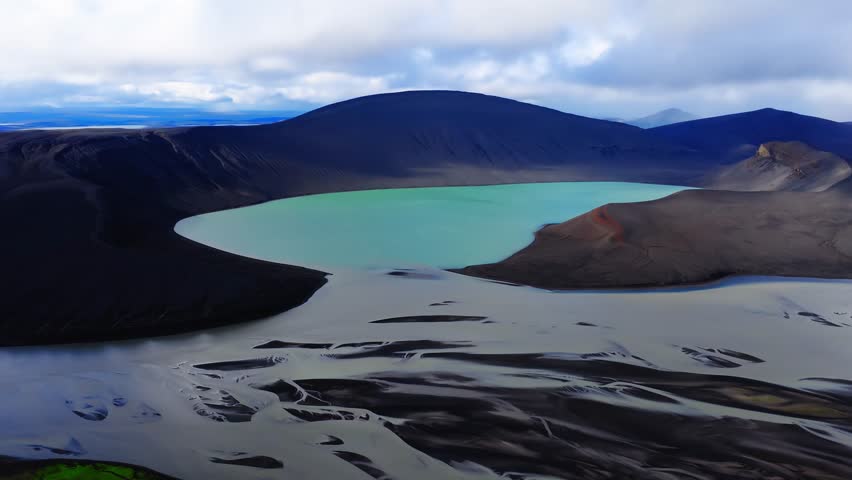 Aerial view shows a milky turquoise crater lake in Iceland in a dark caldera, black slopes, a small reddish cinder cone, and braided outwash on a black sand plain under overcast light.