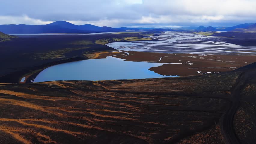 Aerial view shows cobalt lagoon feeding braided meltwater channels across black sand plains, with ochre ridges and volcanic cones under diffuse daylight in Iceland. - Powered by Shutterstock - Get 15% off with code: PIKWIZARD15