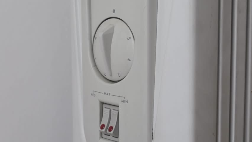 Female hand in a warm green sweater turning the knob of an electric heater to maximum. The heater shows slight signs of wear and dirt - Powered by Shutterstock - Get 15% off with code: PIKWIZARD15
