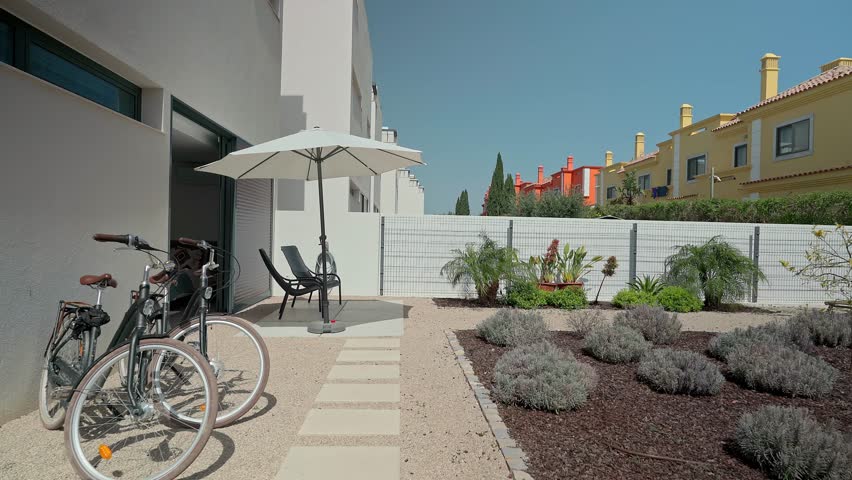 Modern white terraced house backyard with two bicycles parked near a table with chairs under an open umbrella, featuring a small garden with lavender and palm trees
