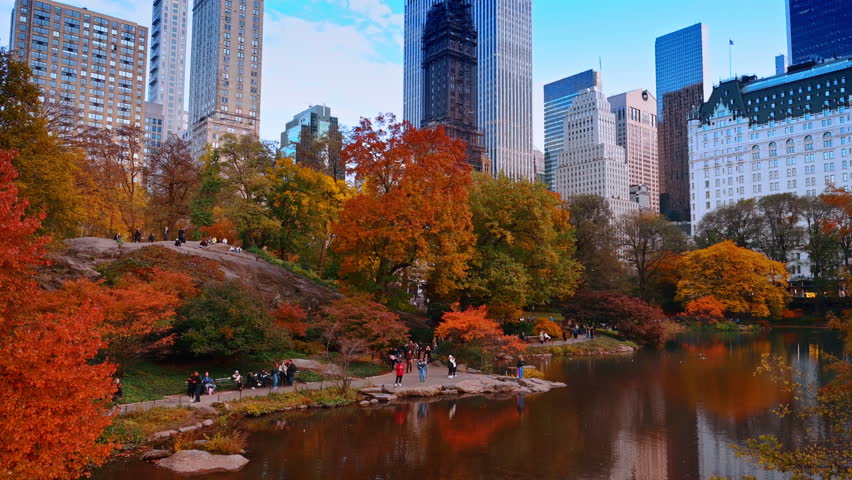 New York, USA, 8 October 2025: Picturesque Autumn at Central Park Lake. People enjoy a peaceful lakeside walk in Central Park surrounded by vivid autumn foliage and reflecting water.