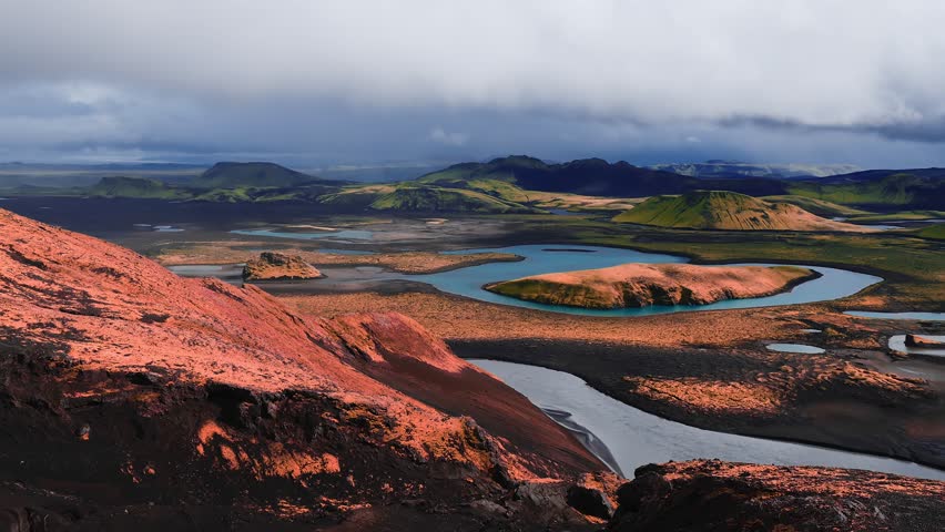 Aerial view of Landmannalaugar in the Icelandic Highlands shows a turquoise braided river, volcanic islands, rhyolite hills, black sand, lava fields, and drifting low clouds.