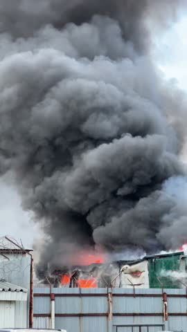 Massive fire engulfing an industrial building, sending thick plumes of black smoke into the sky as firefighters battle the raging blaze