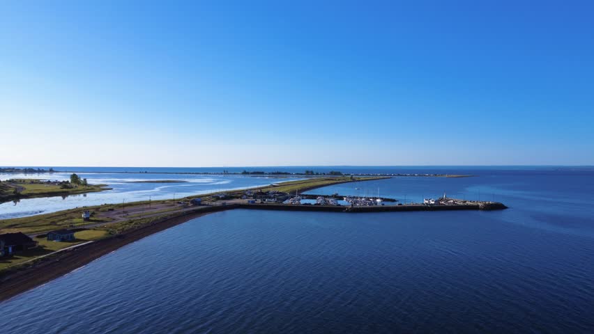 Aerial view of a small marina and its boats, attached to a narrow, verdant peninsula forming a lagoon on a calm, blue sea stretching as far as the eye can see. Carleton-sur-Mer, Quebec, Canada.