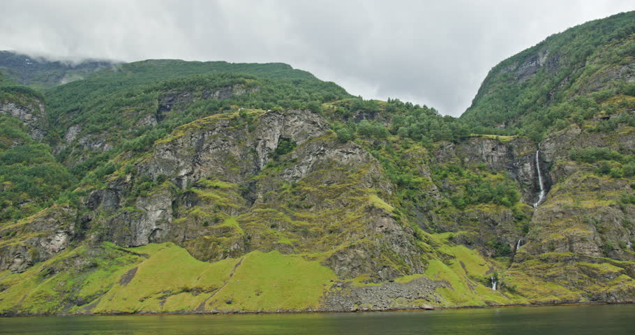 Norway fjord cliffs with lush green slopes and a tall cascading waterfall near Naeroyfjord and Aurlandsfjord, wide scenic view on an overcast day