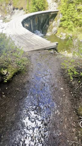POV Walk on a Wooden Boardwalk Trail Over a Melting Alpine Lake and Waterfall in the Mountains