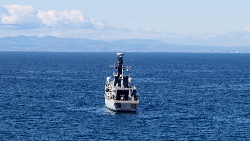 Military vessel sails steadily on blue ocean under clear daylight, distant Scottish coastline visible