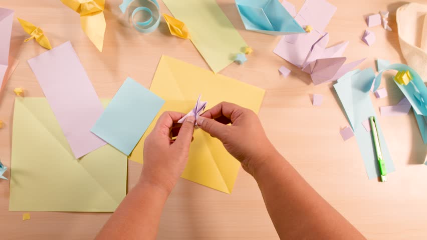 Person folds pastel paper into origami bird, overhead view, bright lighting, colorful craft supplies