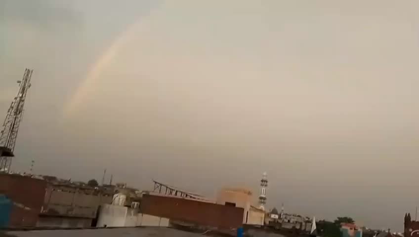 The video shows a colorful rainbow spanning across a cloudy sky over a residential area, likely immediately after a rain shower. A minaret and a communication tower are visible among the buildings.
