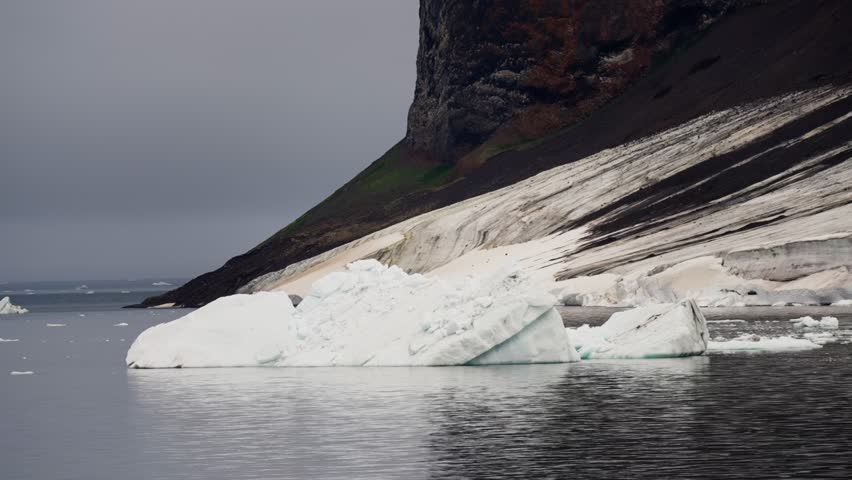 Drifting iceberg against a rugged coastal cliff with layered snow, dark sediment, and calm grey water under an overcast sky, capturing a polar shoreline in a chilly seascape.