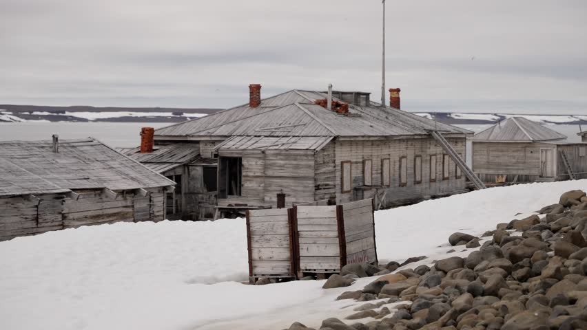 Abandoned wooden outpost on a remote polar coastline, weathered buildings with red-brick chimneys amid melting snow on shore, under an overcast sky at the edge of a frozen sea