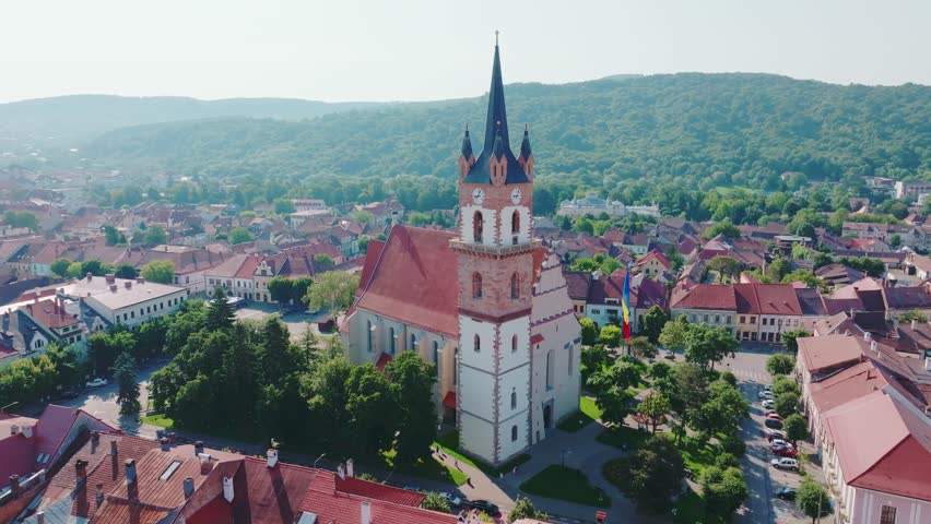 A aerial drone shot performs a slow orbit around the iconic Bistrita Evangelical Church tower, revealing the surrounding historic European city center and distant forested mountains.
