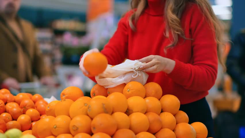 Woman picking oranges into a reusable bag in a store, promoting sustainable shopping, eco-friendly choices, and Earth Day awareness.