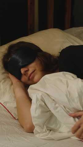 Vertical video. Woman wearing a sleep mask is sleeping in her bed. Person who sleeps peacefully and deeply.